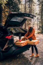 A person standing next to the open trunk of a black Toyota RAV4 which is fitted with a camping conversion kit, featuring a table and sleeping platform inside.